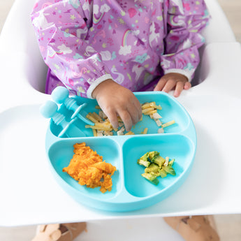 A child wearing a purple bib in a high chair eats veggies & pasta from a Bumkins Silicone Grip Dish: Blue.