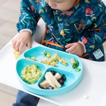 A child in a colorful bib enjoys pasta, fruit, and avocado from a Bumkins Silicone Grip Dish: Blue on a high chair tray.