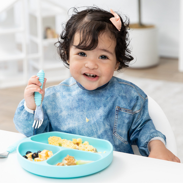 A girl in a blue bib smiles as she eats a meal from Bumkins Grip Dish Essential Toddler 4-Piece Mealtime Set: Blue at a white table.