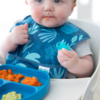 A baby in a Bumkins SuperBib® and Sleeved Bib 3-Piece Set: Hangry, Dinosaurs, Blue Tropics eats orange mashed food from a blue plate.