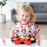 A young girl in a Bumkins Junior Bib: Minnie Mouse Classic smiles at a table with a Minnie Mouse-shaped plate of food and red utensils.