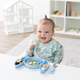 A smiling baby in a pastel bib enjoys fruit from a Bumkins Silicone Grip Dish: Stitch at a white table in a bright playroom.