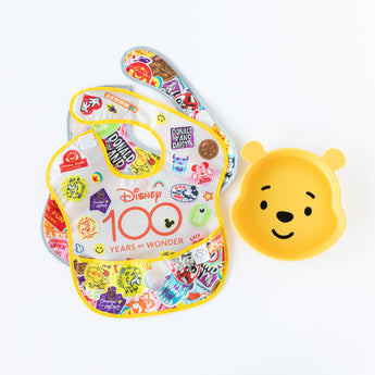 Bumkins Silicone Grip Dish: Winnie The Pooh sits near two overlapping vibrant bibs on a white background.