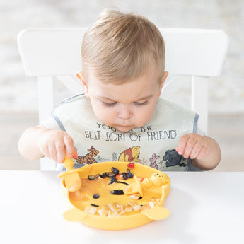 A toddler in a bib eats from a Bumkins Silicone Grip Dish: Winnie The Pooh with matching utensils at a white table.