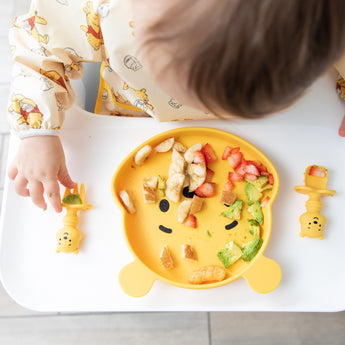 A child enjoys fruit and avocado from a Bumkins Silicone Grip Dish: Winnie The Pooh with matching utensils, seated in a high chair.