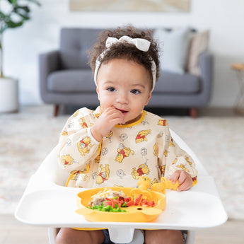 A toddler with a white headband in a high chair enjoys a meal from a Bumkins Silicone Grip Dish: Winnie The Pooh, wearing a yellow bib.