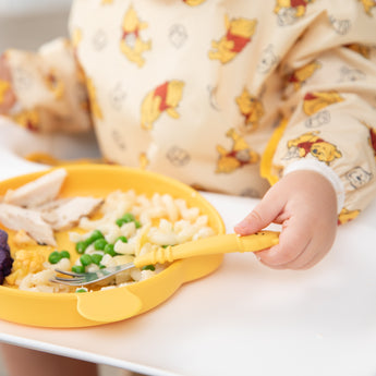 Close-up of a child in a sleeved bib eating with Bumkins Grip Dish Essential 3-Piece Mealtime Set: Disney Winnie the Pooh.