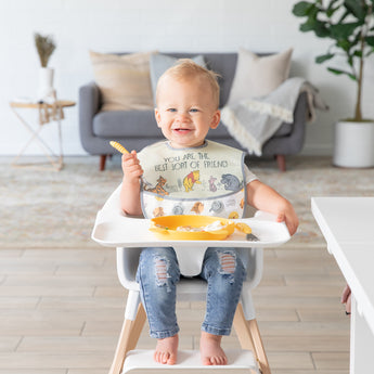 A smiling baby in a high chair, wearing a bib, holds a spoon next to a Bumkins Silicone Grip Dish: Winnie The Pooh in a cozy living room.