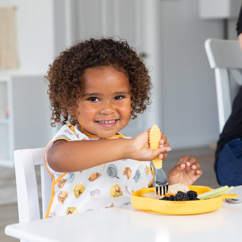 A curly-haired child joyfully eats from a Bumkins Silicone Grip Dish: Winnie The Pooh using a yellow fork at a white table.