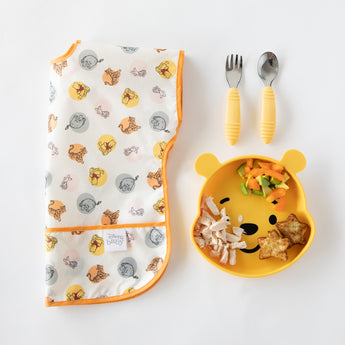 Bumkins Silicone Grip Dish: Winnie The Pooh with chicken and veggies sits near a folded bib and yellow utensils on a white background.