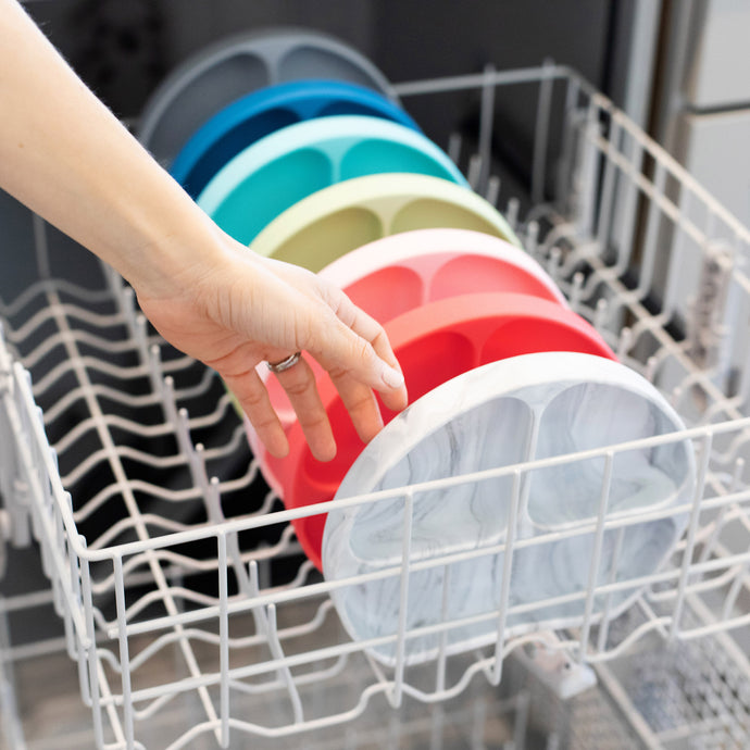 A person places colorful divided silicone plates in a dishwashers top rack. The plates are in shades of grey, blue, teal, green, yellow, red, and pink. The persons hand is visible, with a focus on the orderly arrangement of plates.