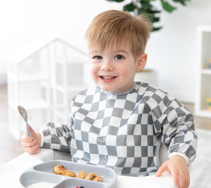 A toddler with light brown hair wearing a gray and white checkered bib sits in a high chair, smiling while holding a spoon. In front of the child is a tray with a sectioned plate containing milk and mini waffles.