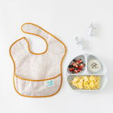 Bumkins Grip Dish Essential Infant 4-Piece Mealtime Set: Marble with fruit, eggs, and yogurt sits by a beige bib on a white background.