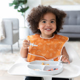 A toddler in a high chair wearing an orange bib eats a meal with Bumkins Grip Dish Essential Infant 4-Piece Mealtime Set: Marble.
