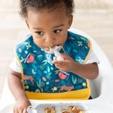 A toddler in a high chair wearing a jungle-themed bib eats from Bumkins Grip Dish Essential Infant 4-Piece Mealtime Set: Marble.