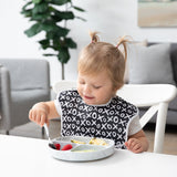 A toddler eats fruit with Bumkins Grip Dish Essential Toddler 4-Piece Mealtime Set: Marble, wearing a black & white bib at a white table.