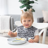 A smiling toddler at a table in a black & white bib enjoys a meal with Bumkins Grip Dish Essential Toddler 4-Piece Mealtime Set: Marble.