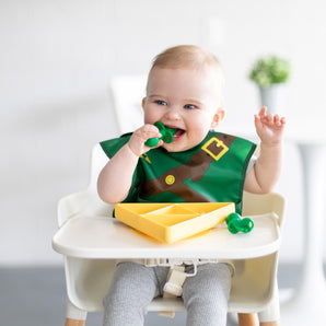 A baby in a high chair wearing a bib, smiling while eating from a Bumkins Silicone Grip Dish: Zelda™ Triforce using green baby utensils.