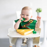 A baby in a high chair wearing a bib, smiling while eating from a Bumkins Silicone Grip Dish: Zelda™ Triforce using green baby utensils.