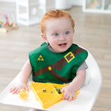 A curly-haired toddler in a high chair, wearing a green bib, eats from a Bumkins Silicone Grip Dish: Zelda™ Triforce on the tray.