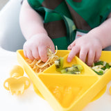 Close-up of a toddler enjoying spaghetti, kiwi, and veggies from a Bumkins Silicone Grip Dish: Zelda™ Triforce on a high chair tray.