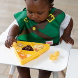 A baby in a high chair enjoys a meal from a Bumkins Silicone Grip Dish: Zelda™ Triforce with yellow utensils.