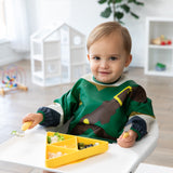 A toddler in a green bib sits in a high chair, gripping a fork and spoon next to a Bumkins Silicone Grip Dish: Zelda™ Triforce on the tray.
