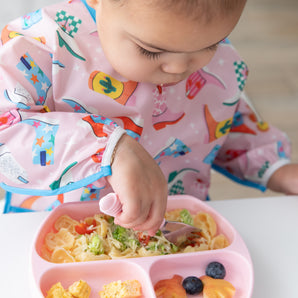 A young girl enjoys self-feeding with Bumkins On-The-Go Mealtime 6-Piece Set: Boots + Pink at a white table.