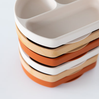 A stack of divided trays in white, light wood, and orange colors, viewed from a slightly elevated angle. Each tray has two compartments. The image focuses on the textures and color contrast of the trays.