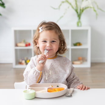 A toddler with a bow clip enjoys a meal from Bumkins Silicone Grip Dish: Sand, securely attached to the white table by its suction base.
