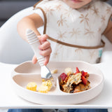Close-up of a child in a high chair using Bumkins Spoon + Fork: Sand to eat from a matching divided plate.