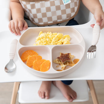 Close-up of a child in high chair holding a fork next to Bumkins Silicone Grip Dish: Sand with scrambled eggs, orange slices & waffles.