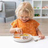 A young blonde boy in an orange bib eats from a tan plate with Bumkins Spoon + Fork: Sand at a white table.