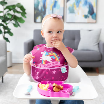 A child in a unicorn bib in a high chair looks curiously to the side as they eat from a Bumkins Silicone Grip Dish Special Edition: Unicorn.