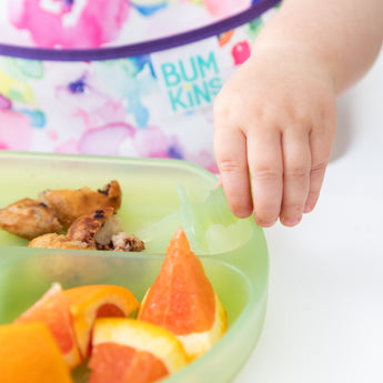 Close-up of a toddler reaching for food from a Bumkins Silicone Grip Dish: Green Jelly, with a colorful bib in the background.