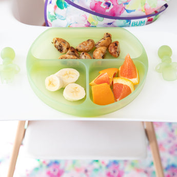 Close-up of a Bumkins Silicone Grip Dish: Green Jelly with bananas, oranges, & pancakes, and green toddler utensils on a high chair tray.
