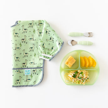 Bumkins Silicone Grip Dish: Green Jelly with cornbread, pasta, and oranges sits next to a llama bib and fork & spoon set on a white background.