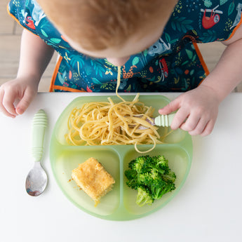 A child with a colorful bib enjoys spaghetti, cornbread, and broccoli from a Bumkins Silicone Grip Dish: Green Jelly at a white table.
