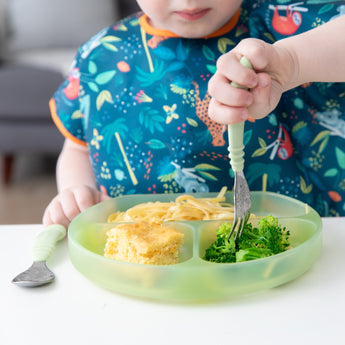 A child enjoys broccoli, pasta, and cornbread from a Bumkins Silicone Grip Dish: Green Jelly, in a vibrant bib.