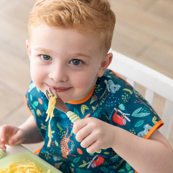 A young child with short red hair smiles while holding a fork with pasta, eating from Bumkins Silicone Grip Dish: Green Jelly.