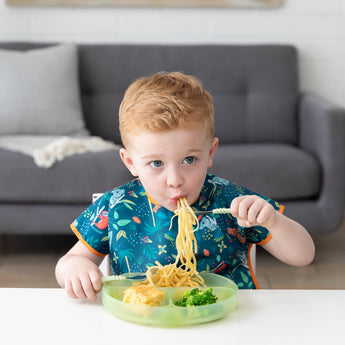 A child in a bib enjoys noodles with a fork, eating pasta and broccoli from a Bumkins Silicone Grip Dish: Green Jelly at a white table.