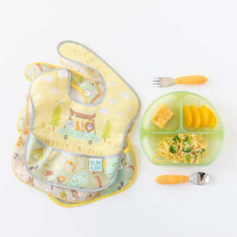 Colorful bibs sit next to Bumkins Silicone Grip Dish: Green Jelly, holding pasta, cornbread, and oranges, and utensils on a white background.