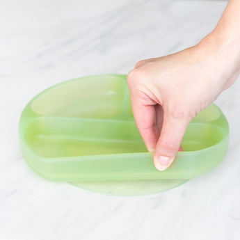 A hand holds a Bumkins Silicone Grip Dish: Green Jelly, demonstrating the suction cup bottom on a marble surface.