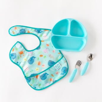 A blue sea-themed bib lays next to Bumkins Grip Dish Essential Toddler 4-Piece Mealtime Set: Blue on a white background.