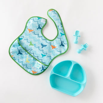 A Bumkins SuperBib® and Sleeved Bib 3-Piece Set: Rolling With The Waves, Whale Tail, Ocean lays by a plate & utensils on a white background.