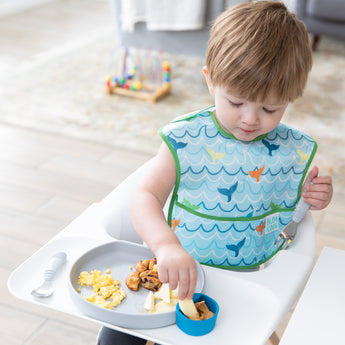 A toddler in a high chair in a Bumkins SuperBib® and Sleeved Bib 3-Piece Set: Rolling With The Waves, Whale Tail, Ocean Life eats from a plate.