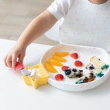 A child in a speckled bib enjoys cake from a Bumkins Silicone Grip Plate: Vanilla Sprinkle, using a red spoon for yogurt in a yellow cup.