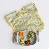 A green bib lays near Bumkins Grip Tray Essential 4-Piece Mealtime Set: Gray with veggies, quinoa, and orange mash on a white background.