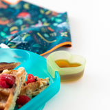 Close-up of a Bumkins Silicone Little Dipper Combo 2 Pack: Aqua Jelly with syrup next to a plate of waffles and a bib on a white background.