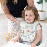 A woman holds Bumkins On-The-Go Mealtime 7-Piece Set: Pooh Bear + Friends as she adjusts a bib on a smiling young girl in a high chair.